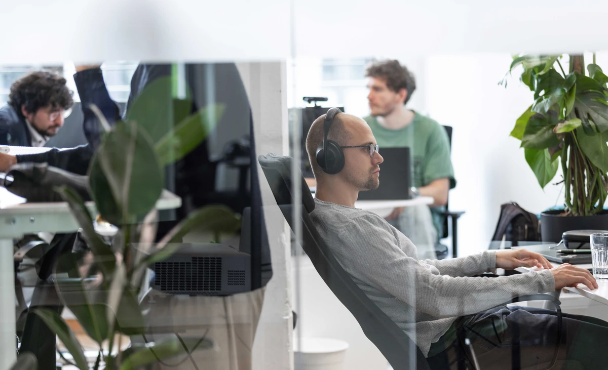 Ein IT-Consultant arbeitet mit Kopfhörern fokussiert an seinem modernen Arbeitsplatz im hellen Kölner Gruppenbüro. Konzentrierte IT Beratung für stabilen Cloud Betrieb